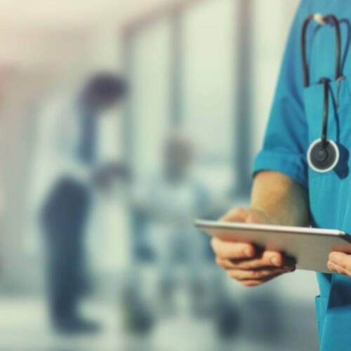 Medical professional wearing blue scrubs with a stethoscope around their neck and holding a tablet device. Blurred background suggests a hospital or clinic setting with staff and patient interaction beyond.
